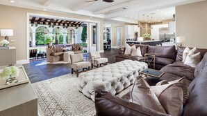 Un salon spacieux avec un grand canapé en cuir marron, un pouf capitonné et un coin repas offrant une vue sur l’extérieur.