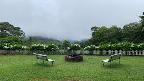 A grassy outdoor area with two benches facing a fire pit, surrounded by a white picket fence and lush greenery. - Villa Chalés Gramado - OH HOTEIS (Gramado)