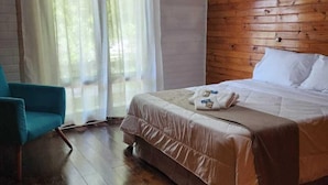 A bedroom with a wooden ceiling, a bed with white linens, a teal chair, and a window with sheer curtains. - Villa Chalés Gramado - OH HOTEIS (Gramado)