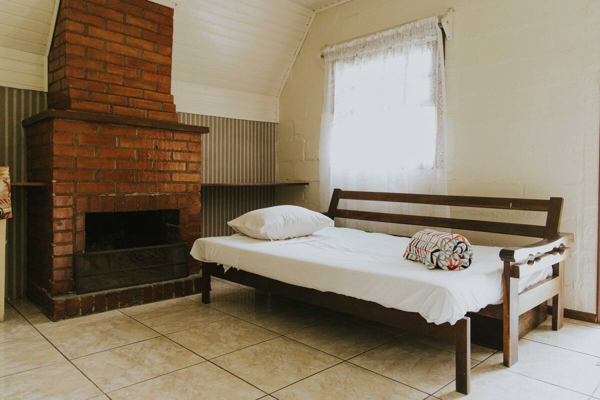 Une chambre avec une cheminée en briques, un lit en bois et une fenêtre ornée d’un rideau blanc.