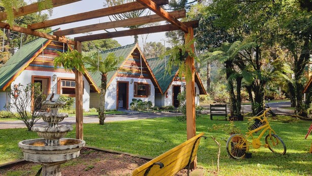 A garden with a wooden pergola, a fountain, and a yellow bicycle. - Villa Chalés Gramado - OH HOTEIS (Gramado)