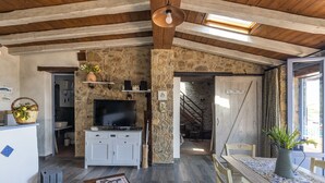 Un interior rústico con pared de piedra, vigas de madera, televisor y una escalera que sube a un nivel superior.