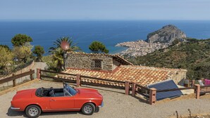 Un descapotable rojo estacionado frente a un edificio de piedra con techo de tejas, con vistas a un pueblo costero y una montaña.