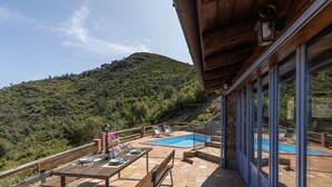 Una terraza con piscina, mesas y sillas de madera, con vistas a una colina.