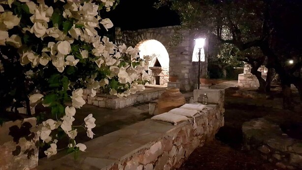 Ein Kiesweg führt zu einem rustikalen Steinhaus mit rundem Bogenportal, umgeben von weißen Blumen und Steinmauern.