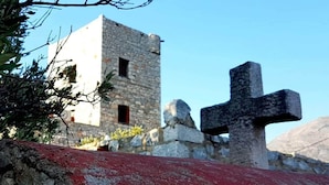 Ein steinernes Gebäude mit Kruzifix davor, umgeben von felsigem Gelände und vereinzelter Vegetation.