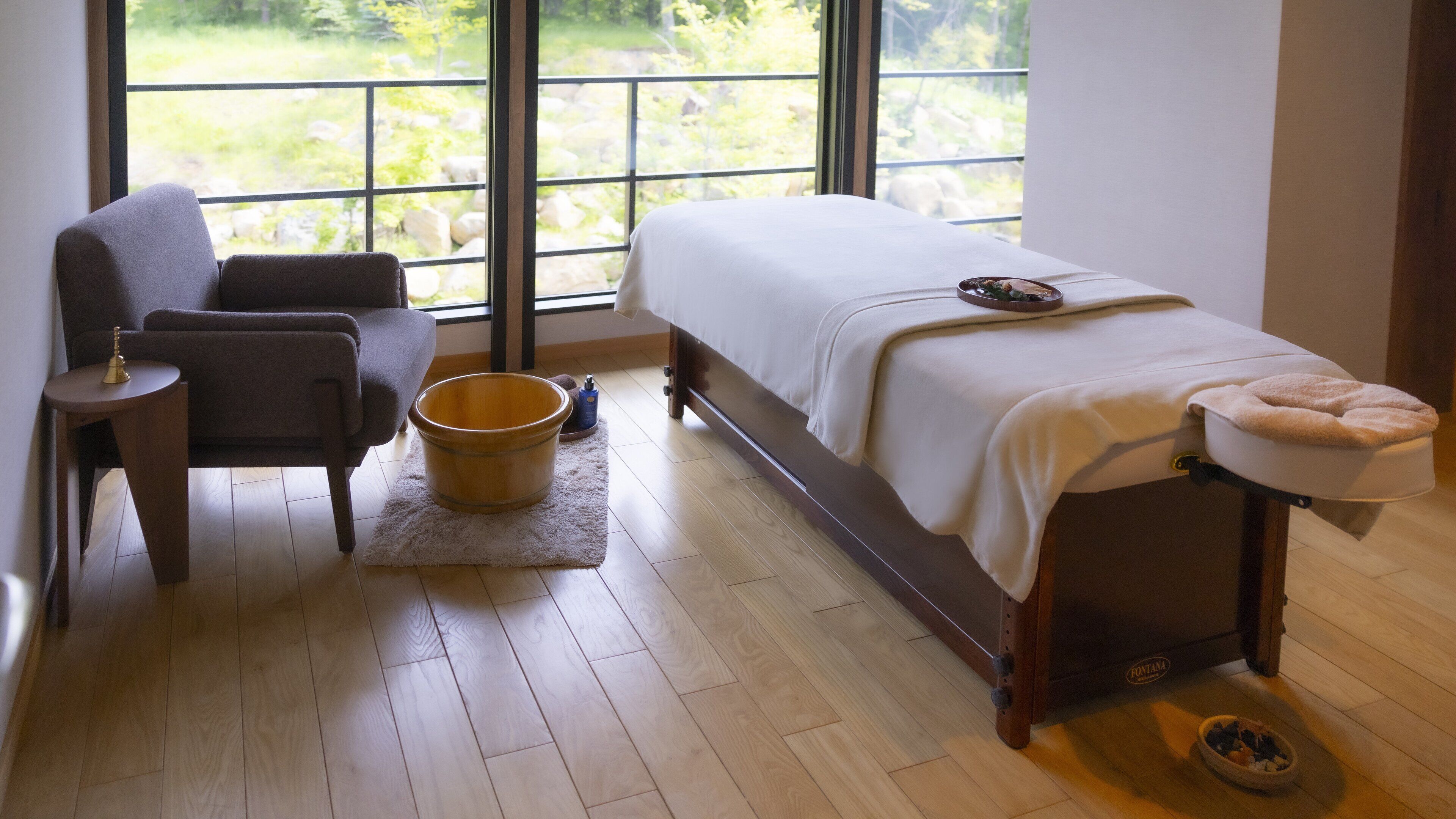 Una stanza con un letto, una sedia, un catino di legno, un piccolo tappeto e una finestra vista verde.