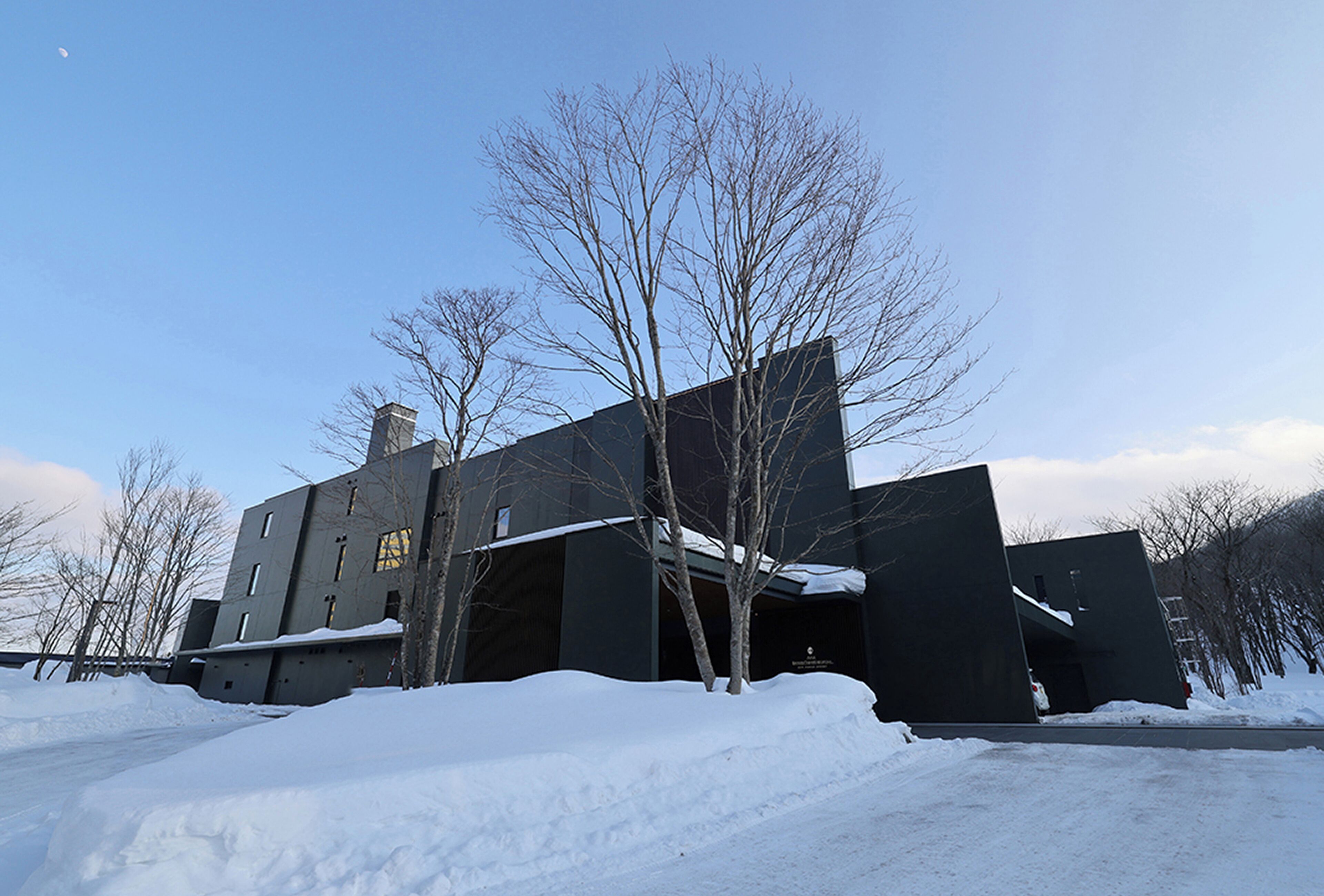 Un edificio grande y oscuro con techo plano y chimenea, rodeado de terreno cubierto de nieve y árboles sin hojas.