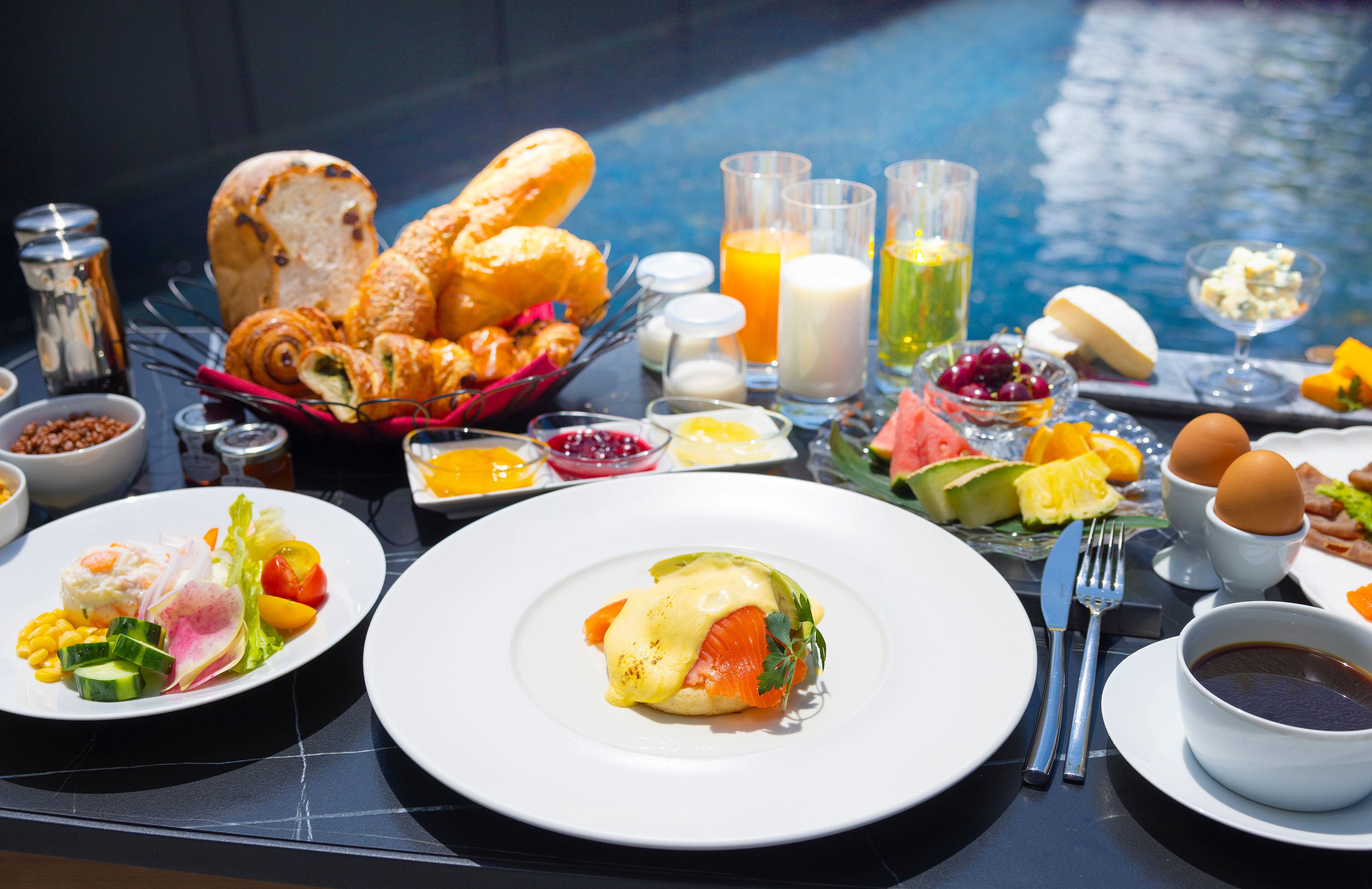 A breakfast buffet with various dishes, including eggs, pastries, and fruits, set against a backdrop of a swimming pool.