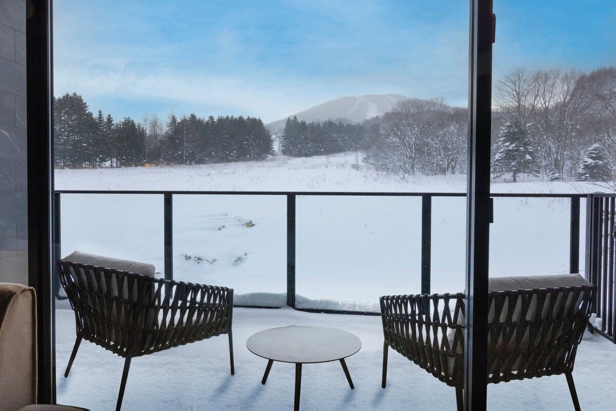 A balcony with two wicker chairs and a small table, overlooking a snowy landscape with trees and a mountain.
