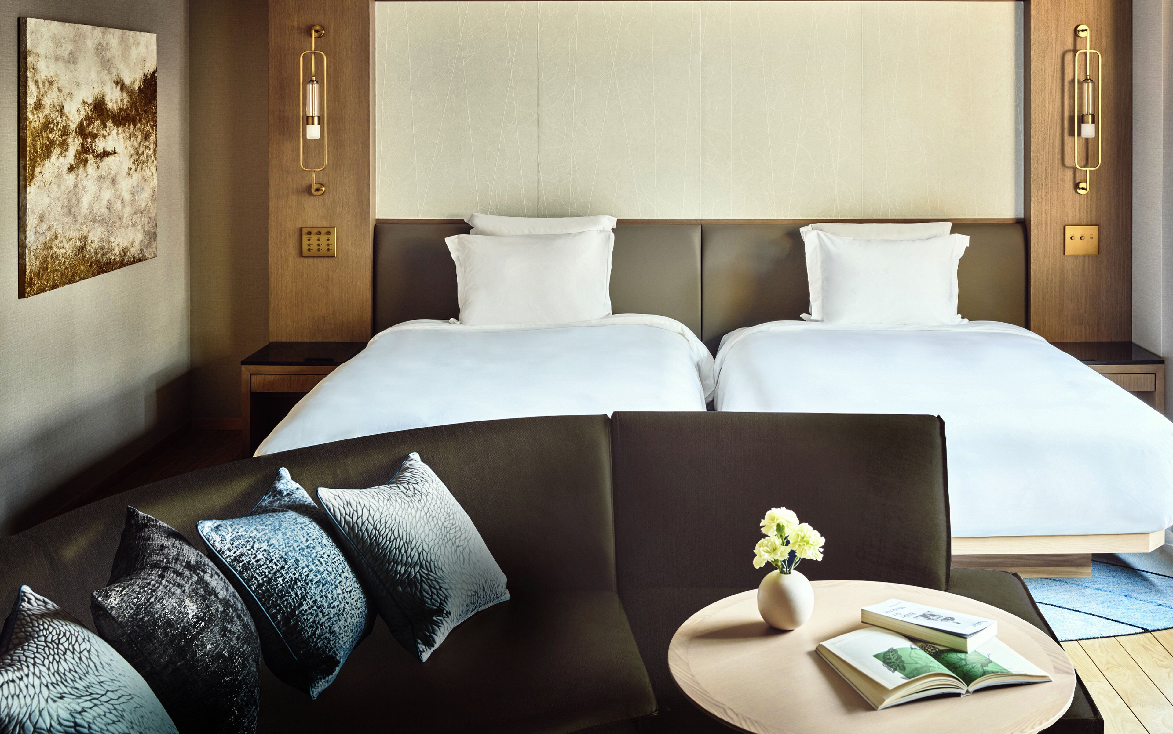 Habitación de hotel con cama doble, sofá, una pequeña mesa con un florero y un libro, y una lámpara de pared.
