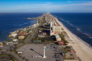 An aerial view of a coastal city with a prominent beach, a large parking area, and a tall building near the shore.