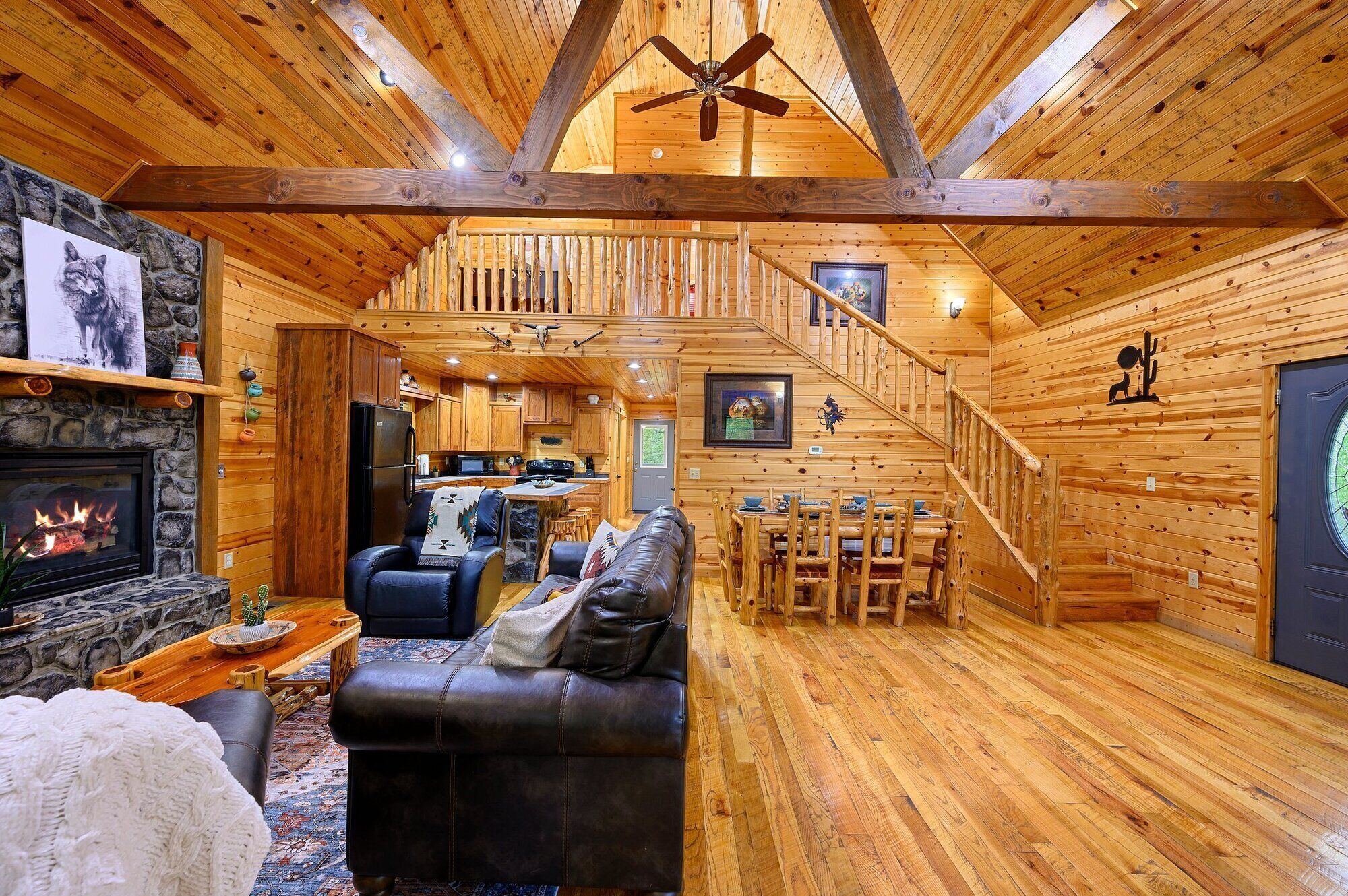 A cozy log cabin interior with a stone fireplace, leather sofa, dining area, and a staircase leading to an upper level.