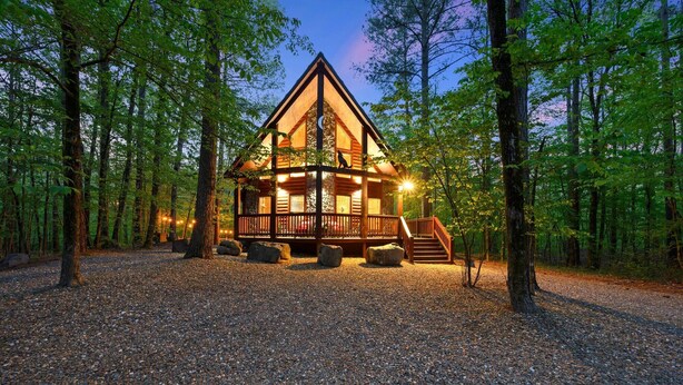 A log cabin with a wooden deck and a staircase leading to the entrance, surrounded by a forest.