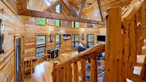 A wooden cabin interior with a high ceiling, wooden beams, and a staircase leading to a loft area.