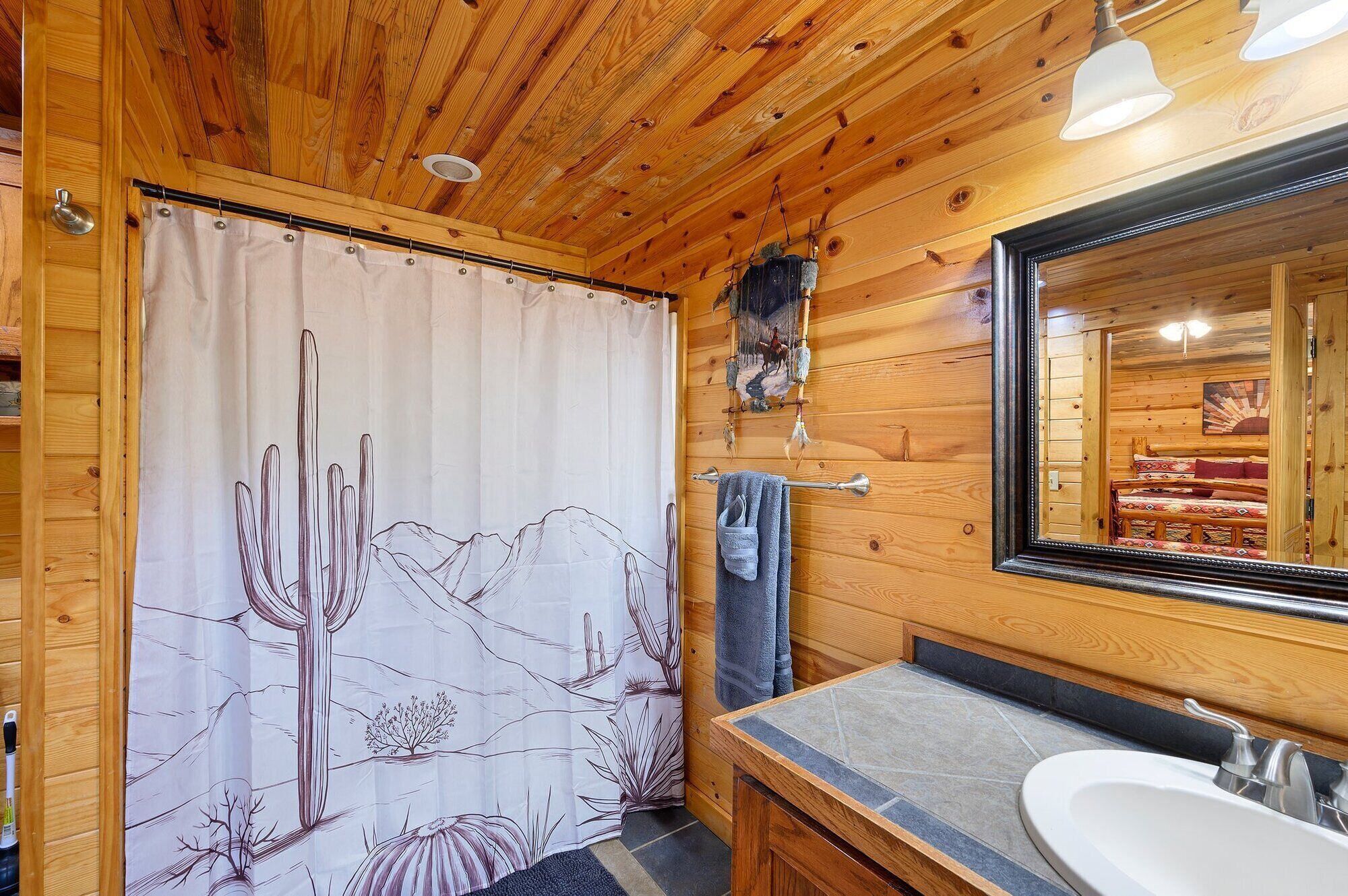 A wooden cabin bathroom with a shower curtain featuring a desert landscape, a large mirror, and a sink with a stone countertop.