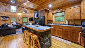 A cozy cabin interior with a stone island bar, wooden cabinets, a fireplace, and a large window overlooking greenery.