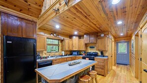 A spacious kitchen with wooden cabinets, a black refrigerator, a central island with stools, and a view of the outdoors through the window.
