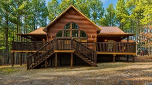 Une cabane en rondins avec un escalier en bois menant à une terrasse, le tout entouré d’arbres.