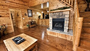 L’intérieur d’une cabane en rondins, avec une cheminée en pierre, un plancher en bois, un coin repas et un escalier.