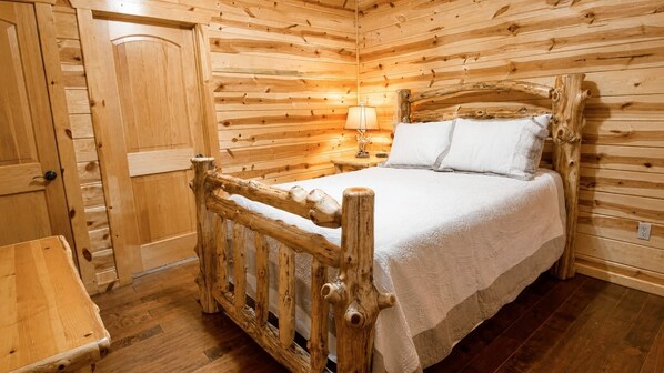 A wooden cabin room with a bed, a nightstand, and a lamp. - Juniper Haven | 2BR + Loft, Hot Tub & Pets (Broken Bow)