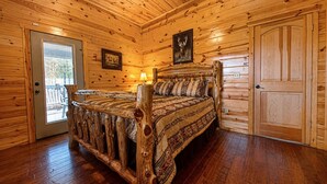 A log cabin bedroom with a wooden bed, a door, and a window. - Juniper Haven | 2BR + Loft, Hot Tub & Pets (Broken Bow)