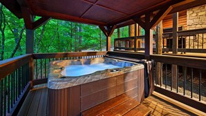 Ein Whirlpool auf einer Holzterrasse mit einer Steinmauer und Holzgeländer, umgeben von Bäumen.