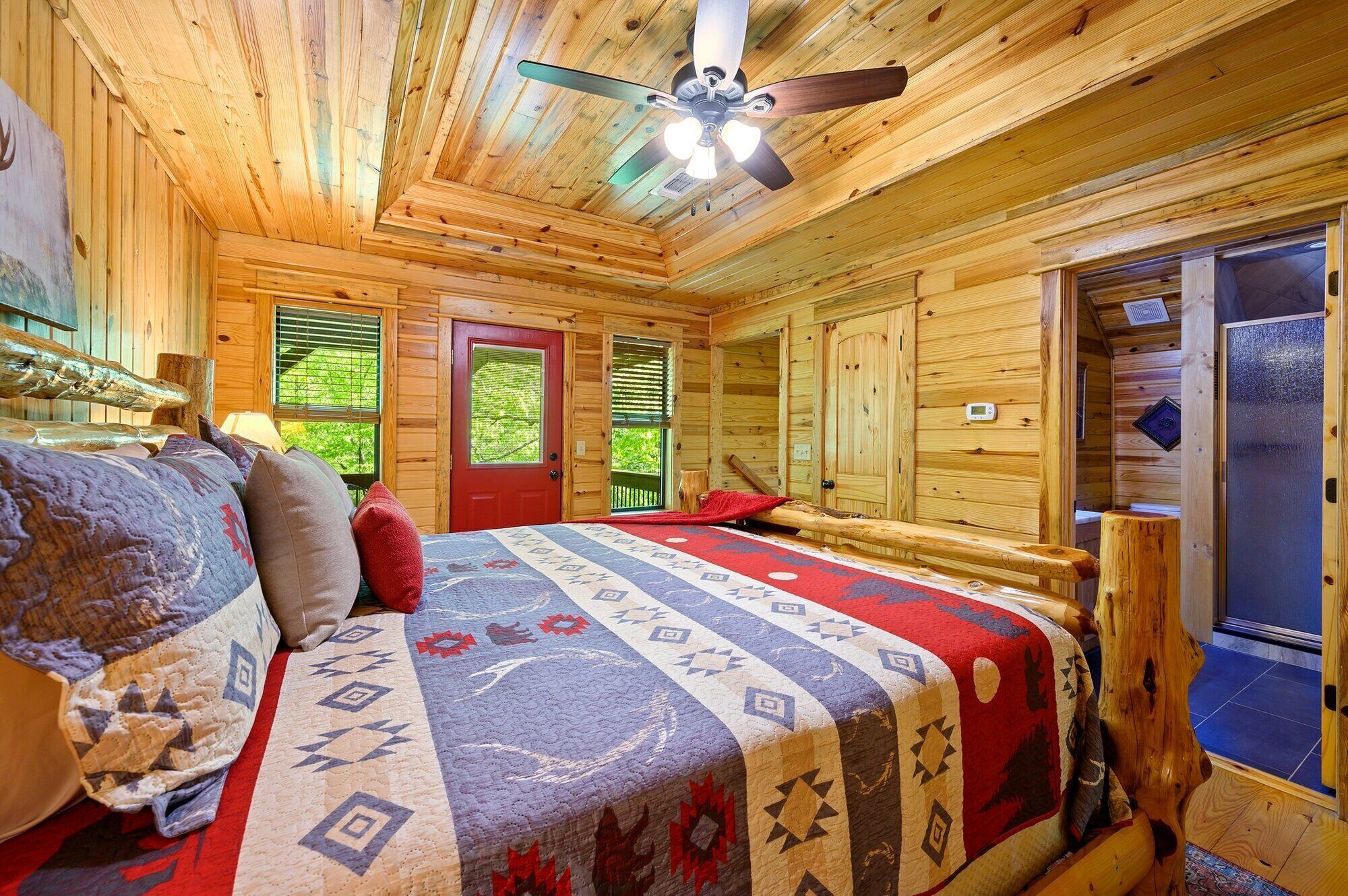 A cozy log cabin room with a bed, a ceiling fan, and a window with a view of greenery.