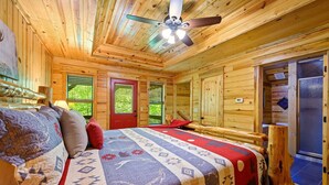 Ein gemütliches Blockhauszimmer mit Bett, Deckenventilator und Fenster mit Blick auf Grünflächen.