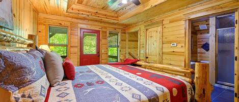 A cozy log cabin room with a bed, a ceiling fan, and a window with a view of greenery.