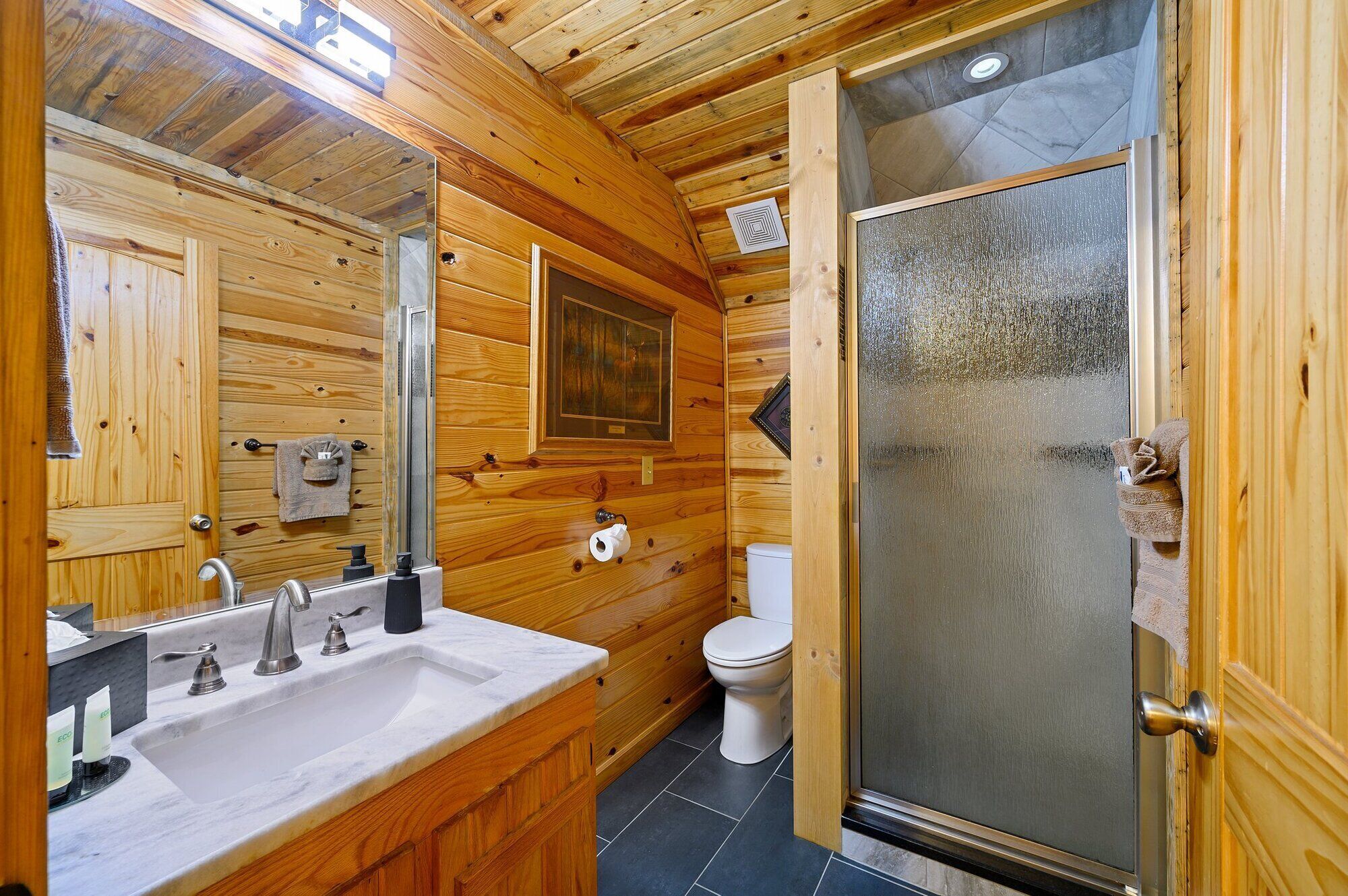 A wooden cabin bathroom with a large mirror, a sink with a marble countertop, a toilet, and a glass-enclosed shower.