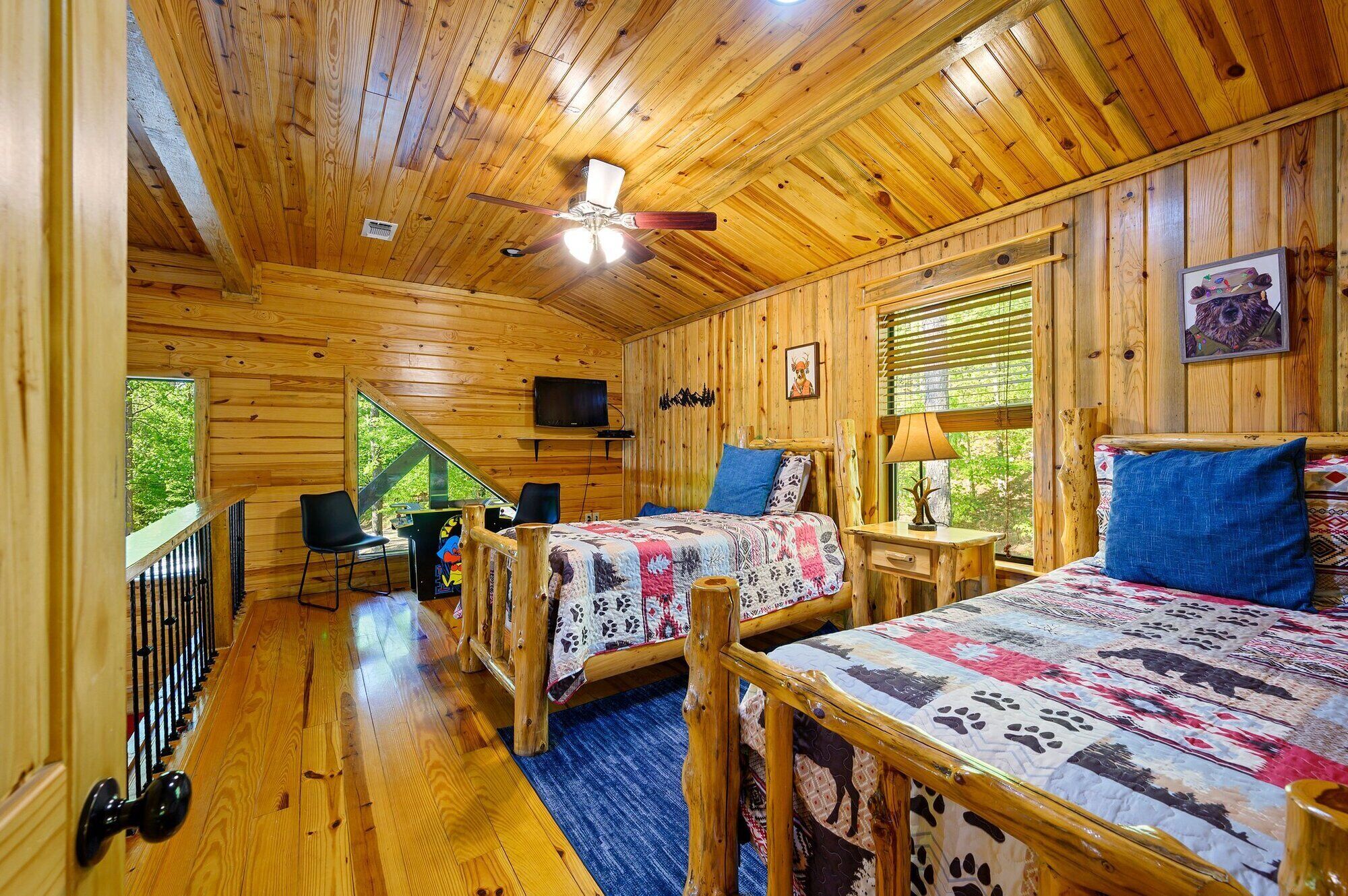 Une cabane comprenant deux chambres, chacune étant équipée d’un lit, d’un bureau et d’une chaise. Un téléviseur est fixé au mur, et une fenêtre est dotée de stores.