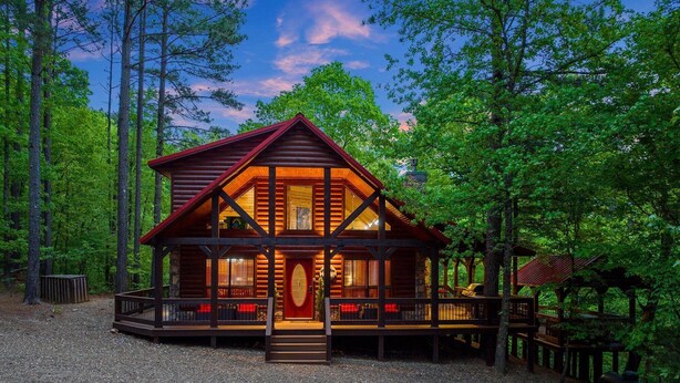 Eine Blockhütte mit Veranda und roter Tür, umgeben von Bäumen.