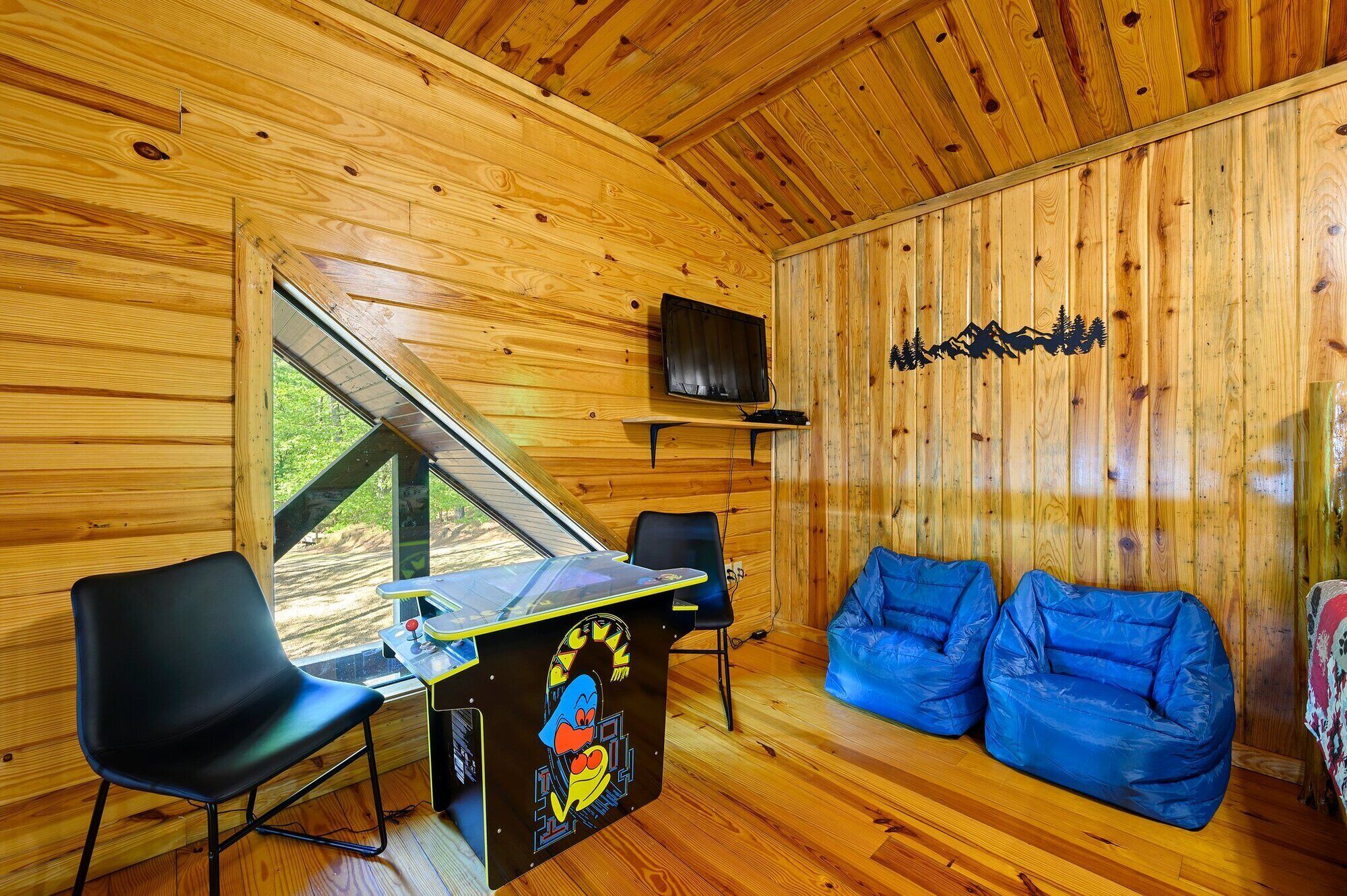Interior de una cabaña de madera con un escritorio, sillas, un sofá azul y un televisor montado.