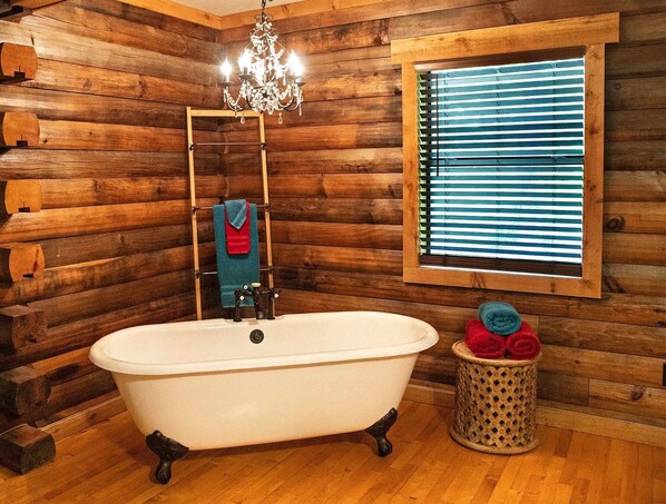 A log cabin bathroom with a clawfoot bathtub, a wooden ladder, a chandelier, and a window with blinds.