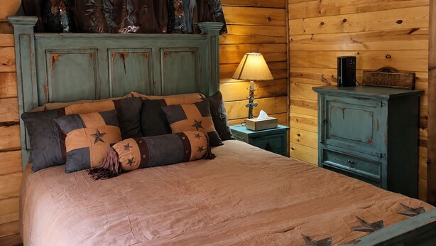 Een bed met een beige dekbedovertrek en kussens met een sterrenpatroon, een nachtkastje met een lamp en een houten kastje met een bordje.