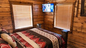 Une chambre dans un chalet en bois, avec un lit, deux fenêtres équipées de stores, un téléviseur fixé au mur et une petite table.