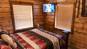 Een houten kamer met een bed, twee ramen met zonwering, een televisie aan de muur en een kleine tafel.