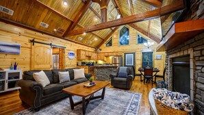 A spacious living room with a fireplace, wooden ceiling beams, a stone accent wall, and a cozy seating area with a couch and armchairs. - Logan's Bluff Cabin at Hochatown (Broken Bow)