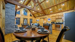 A wooden cabin interior with a dining table set for four, a kitchen with modern appliances, and a stone wall. - Logan's Bluff Cabin at Hochatown (Broken Bow)