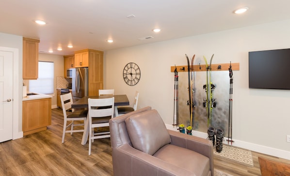 Une cuisine moderne avec des armoires en bois et un réfrigérateur en acier inoxydable. On y trouve un coin repas avec une table et des chaises, ainsi qu’une grande horloge murale. Du matériel de ski est fixé au mur.