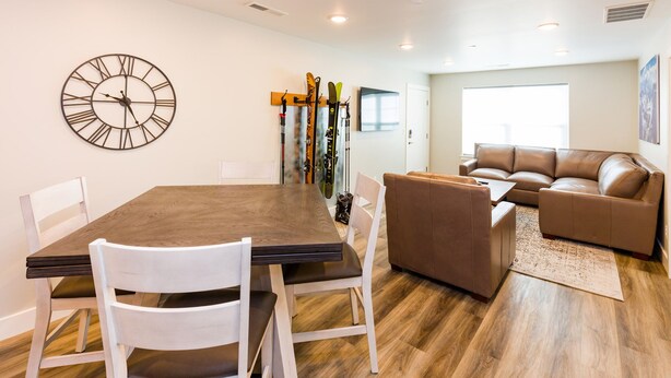 A modern living room with a wooden dining table, white chairs, a brown leather sofa, and a large wall clock.