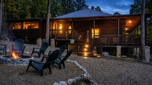 A rustic cabin with a fire pit and outdoor seating, surrounded by a gravel area and trees.