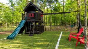Een houten speelconstructie met een glijbaan en schommelset in een gazon, omringd door bomen.