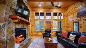 A cozy log cabin interior with a stone fireplace, leather sofa, and a television mounted on the wall.