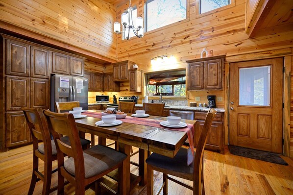 A wooden cabin-style kitchen with a dining table set for four, wooden cabinets, and a large window. - The Kootenai Creek Cabin at Hochatown (Broken Bow)
