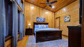 A bedroom with wooden walls, a bed with blue bedding, a ceiling fan, and a large rug.