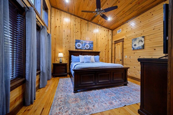 Un dormitorio con paredes de madera, una cama con ropa de cama azul, un ventilador de techo y una alfombra grande.