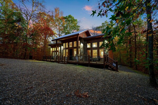 A log cabin with a deck, surrounded by trees and gravel.