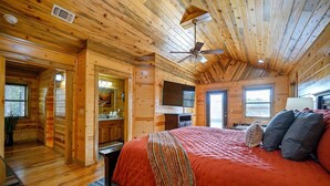 Un dormitorio tipo cabaña de madera con una cama grande, ventilador de techo, televisor y un baño con armarios de madera.